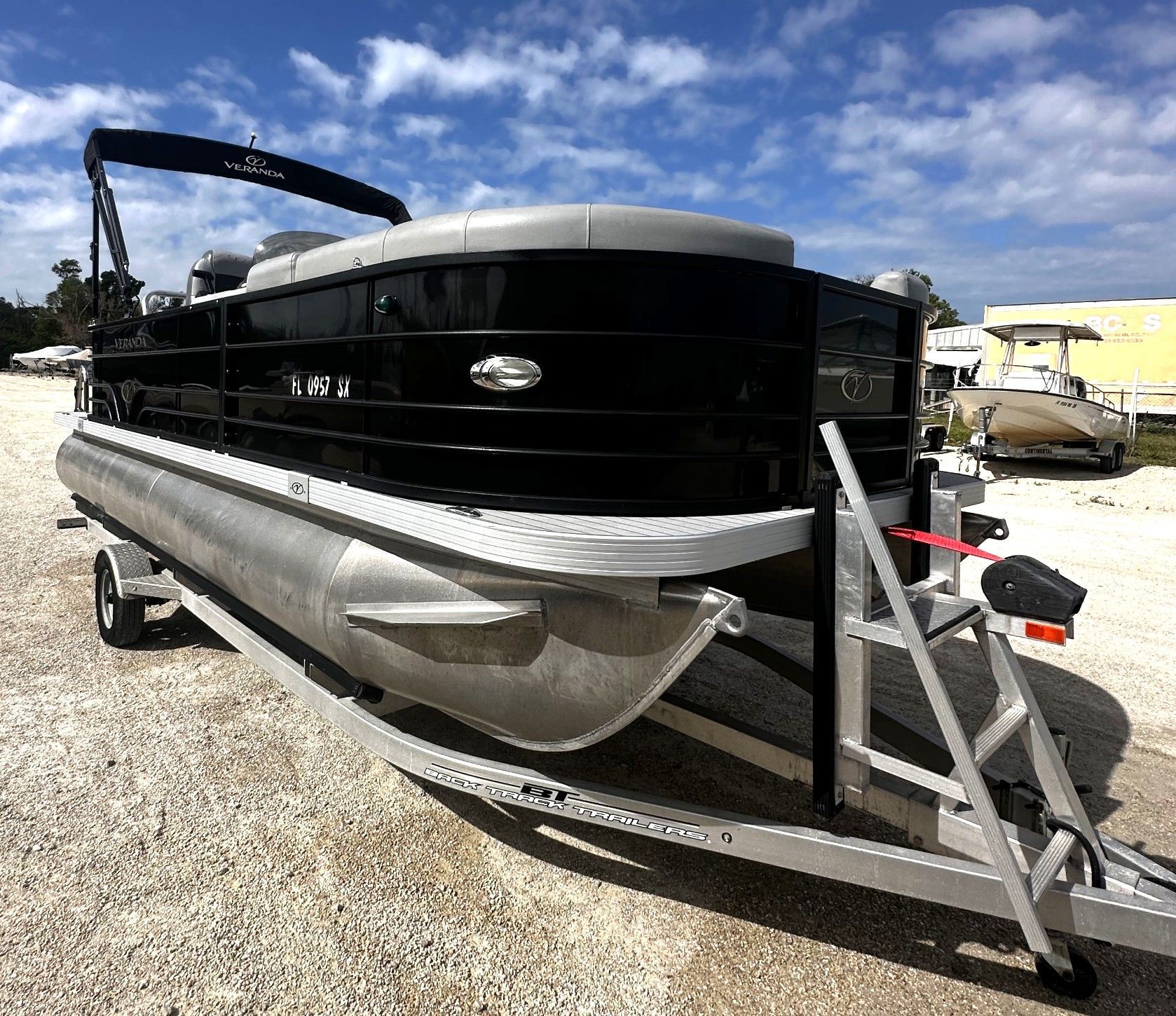 Black and silver pontoon boat on a trailer in a gravel lot, with a dock ladder attached
