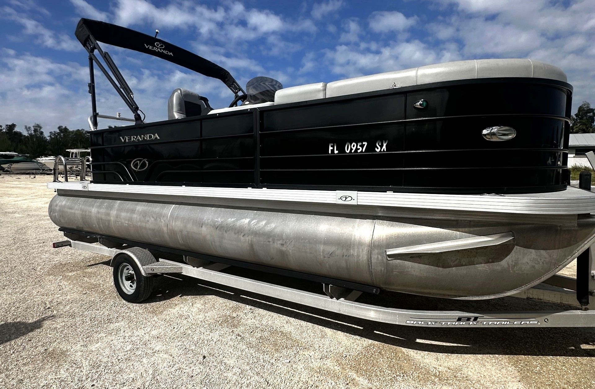 Black pontoon boat on a trailer in a gravel lot under a blue sky.