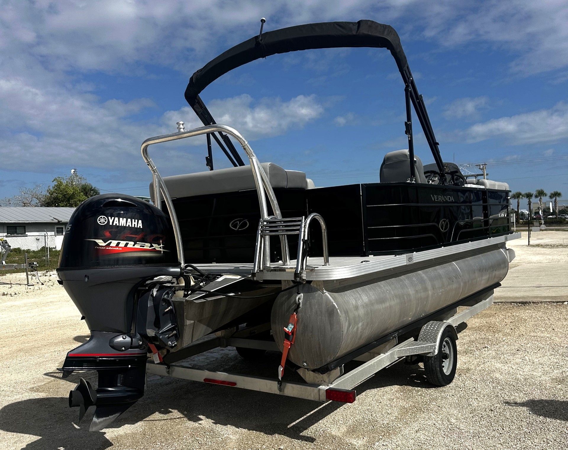 Pontoon boat on a trailer with black canopy and outboard motor, parked on a sunny beach.