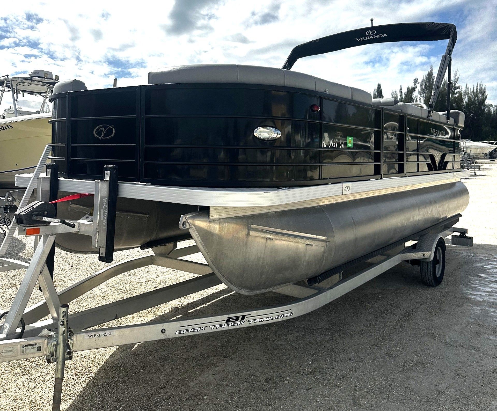 Black pontoon boat on trailer parked outdoors by the water under an overcast sky.