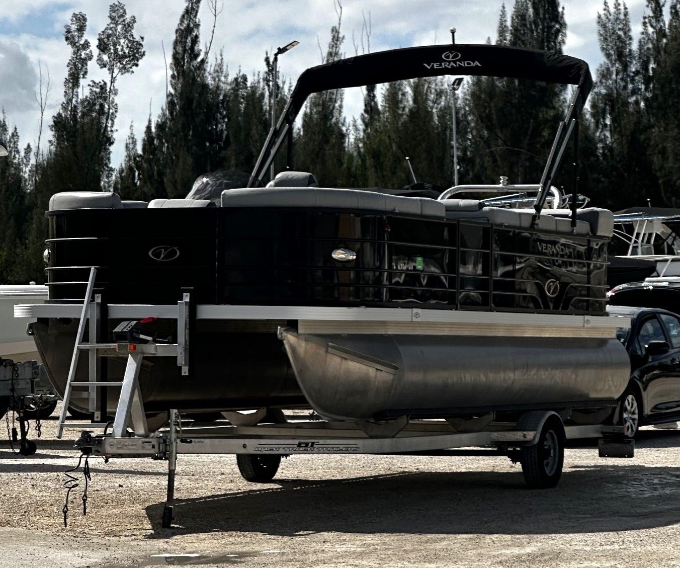 Black pontoon boat on a trailer parked outdoors with trees in the background