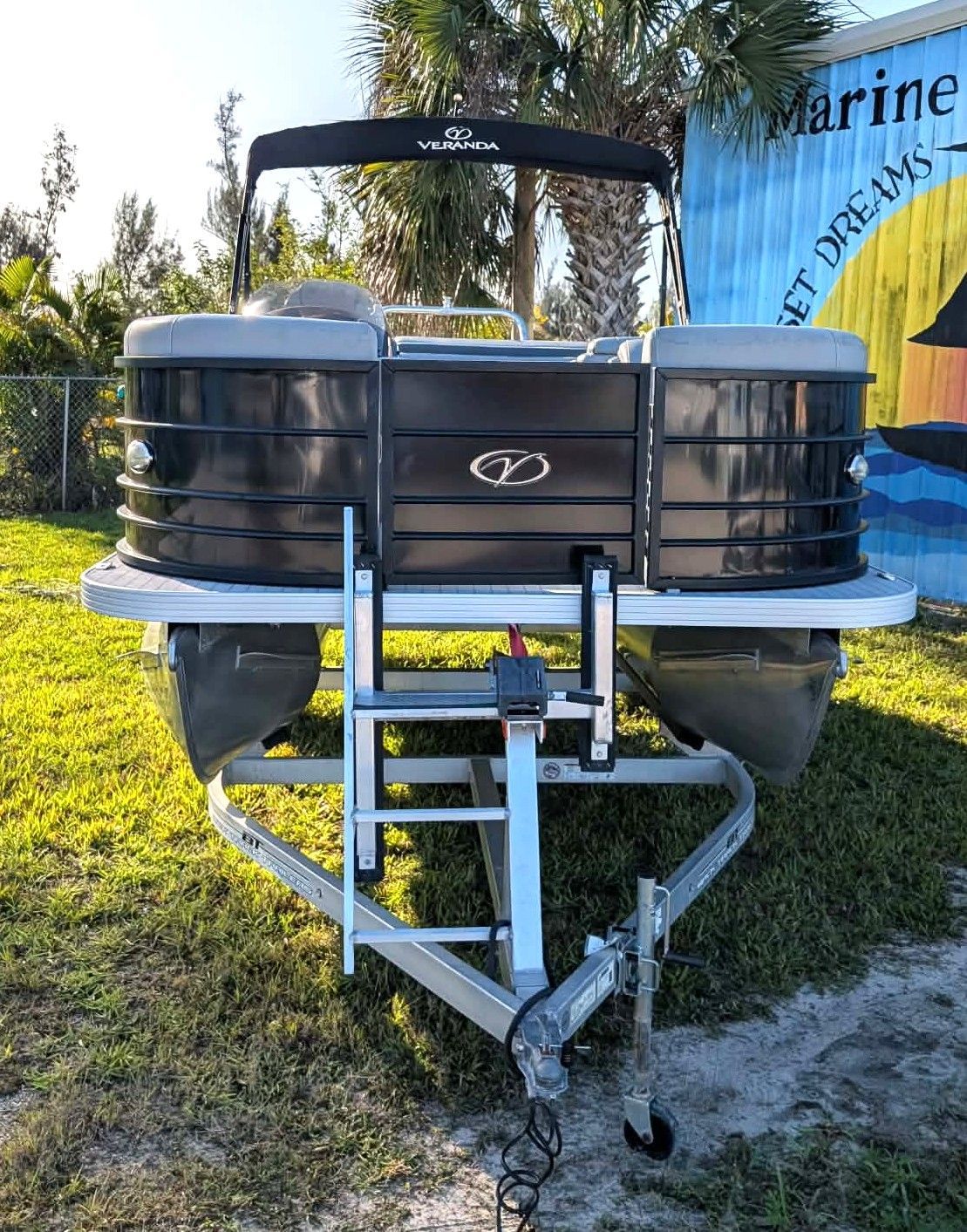 Silver pontoon boat front on a trailer in a grassy yard, with a canopy and blue marine banner behind.