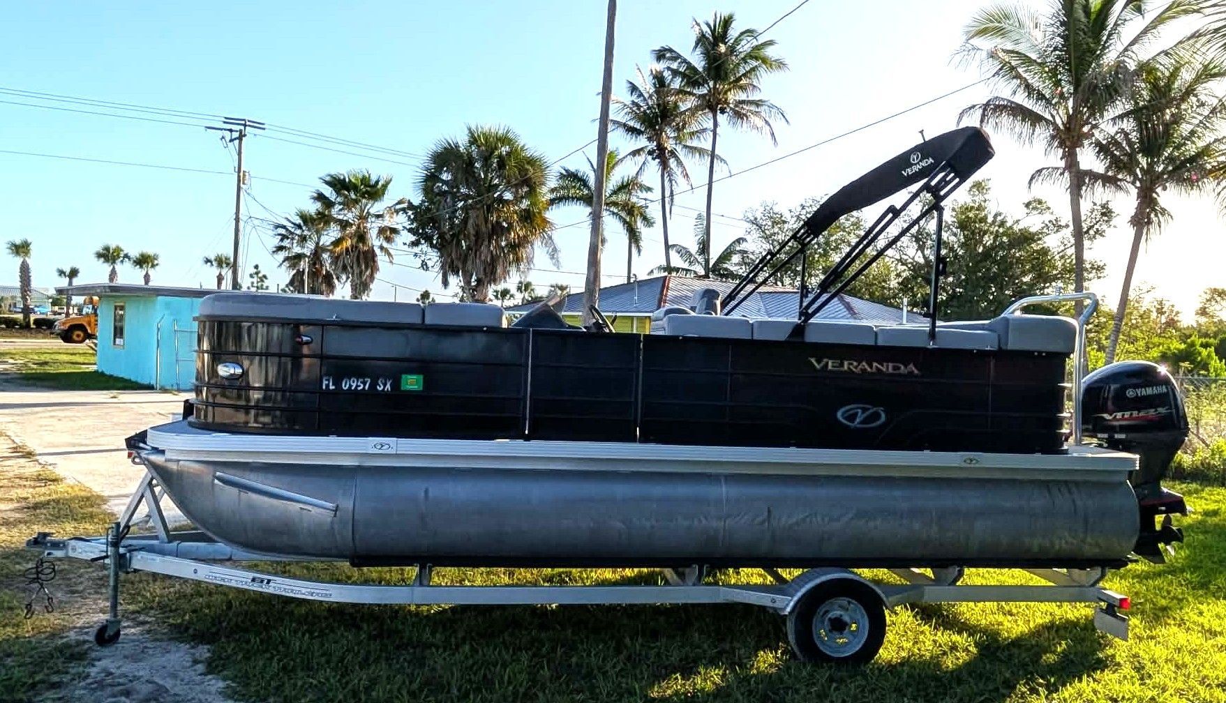 Pontoon boat on a trailer in a grassy lot, with palm trees and water in the background