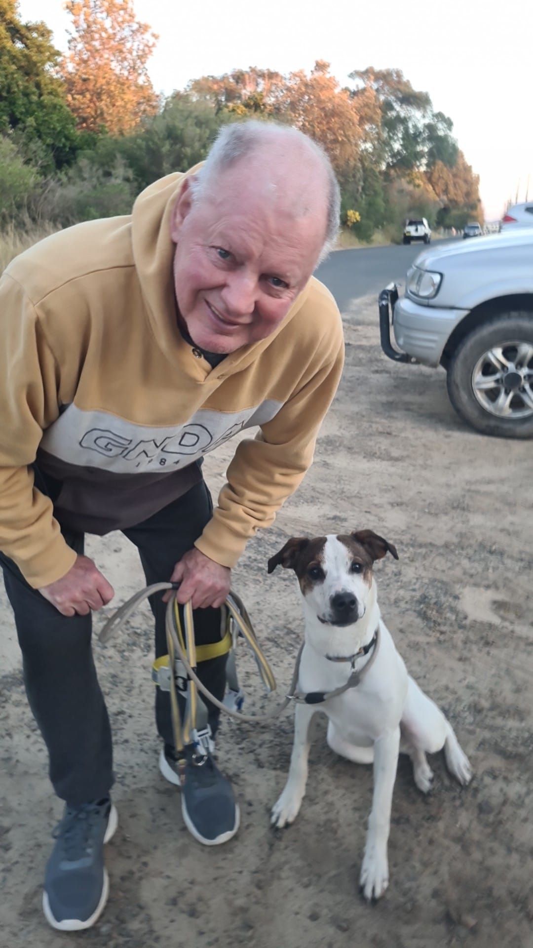 Man with prosthetic leg smiles next to a small dog on a dirt path. Car and trees in the background — Amanda's Professional K9 Training In Forster, NSW