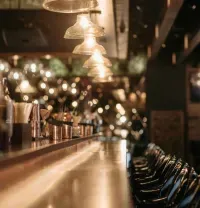 An empty bar counter with warm, glowing pendant lights, wood surfaces, and a row of dark stools in a dim, upscale setting.