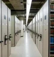A long, narrow aisle between rows of tall, light-colored mobile library shelving units with hand cranks on a tile floor.