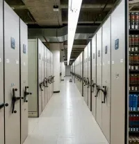 A long, narrow aisle between rows of tall, light-colored mobile library shelving units with hand cranks on a tile floor.