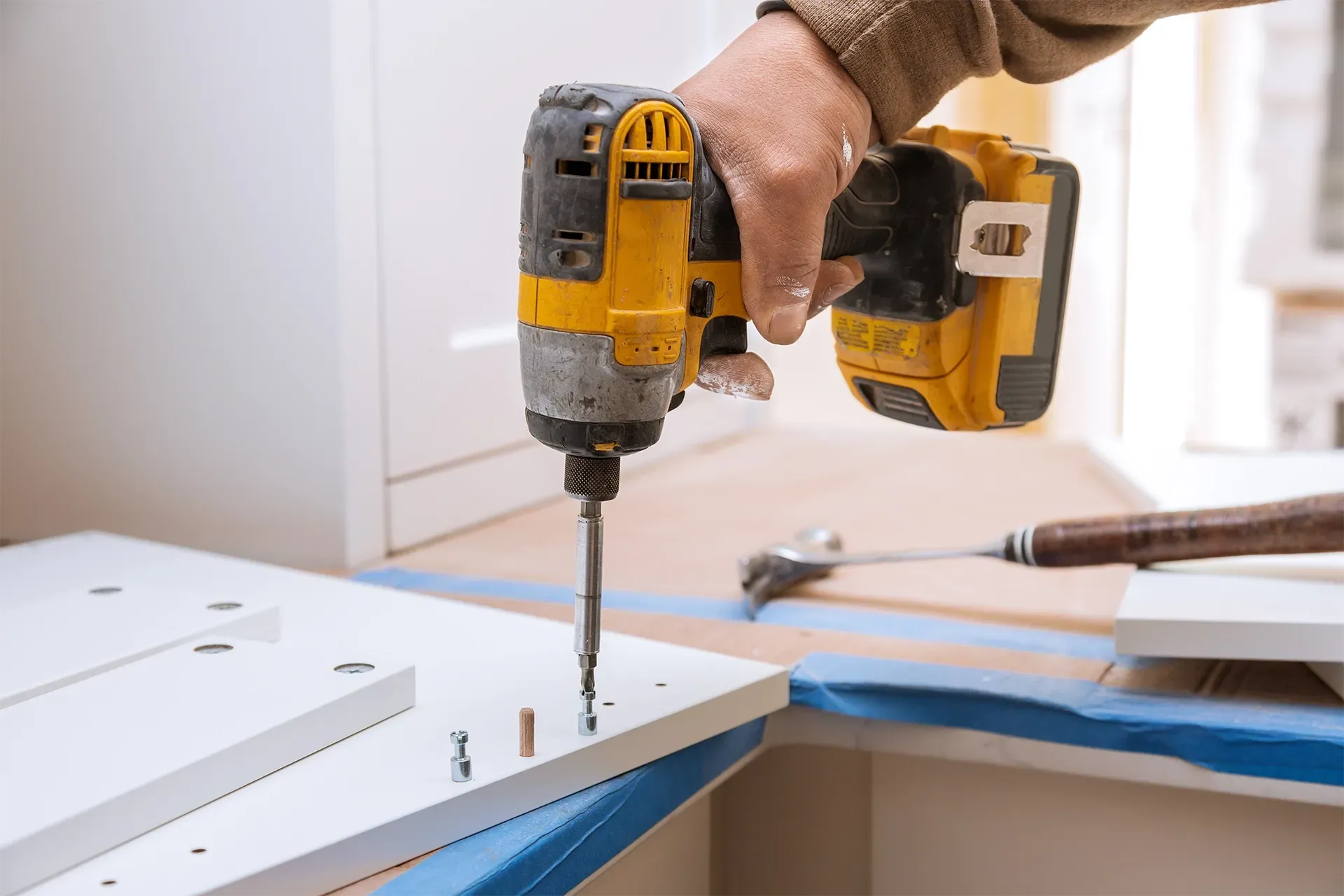 A hand uses a yellow cordless power drill to drive a screw into a white flat-pack furniture panel.