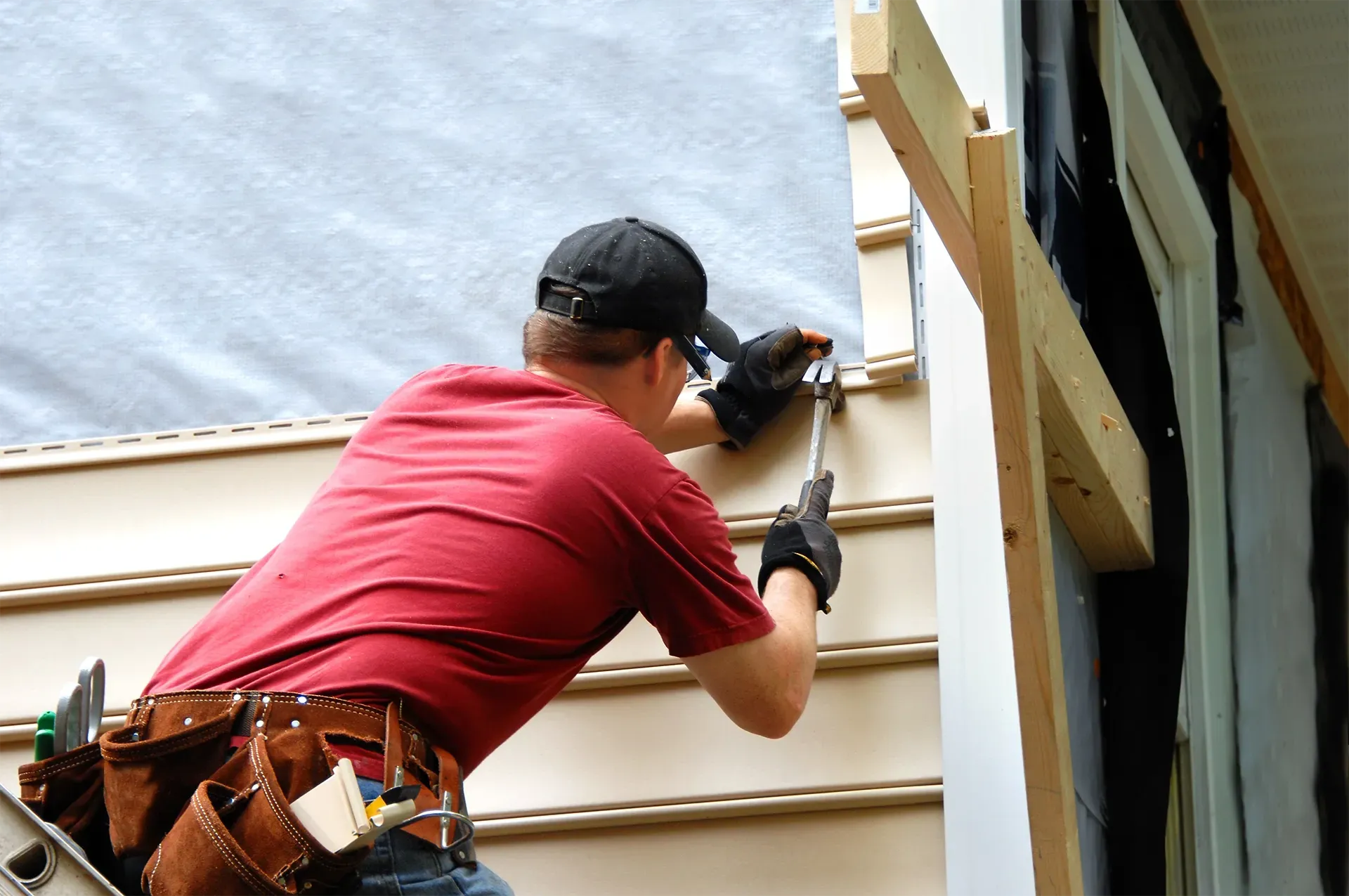 A worker in a red shirt and tool belt installs beige vinyl siding on the exterior wall of a building.