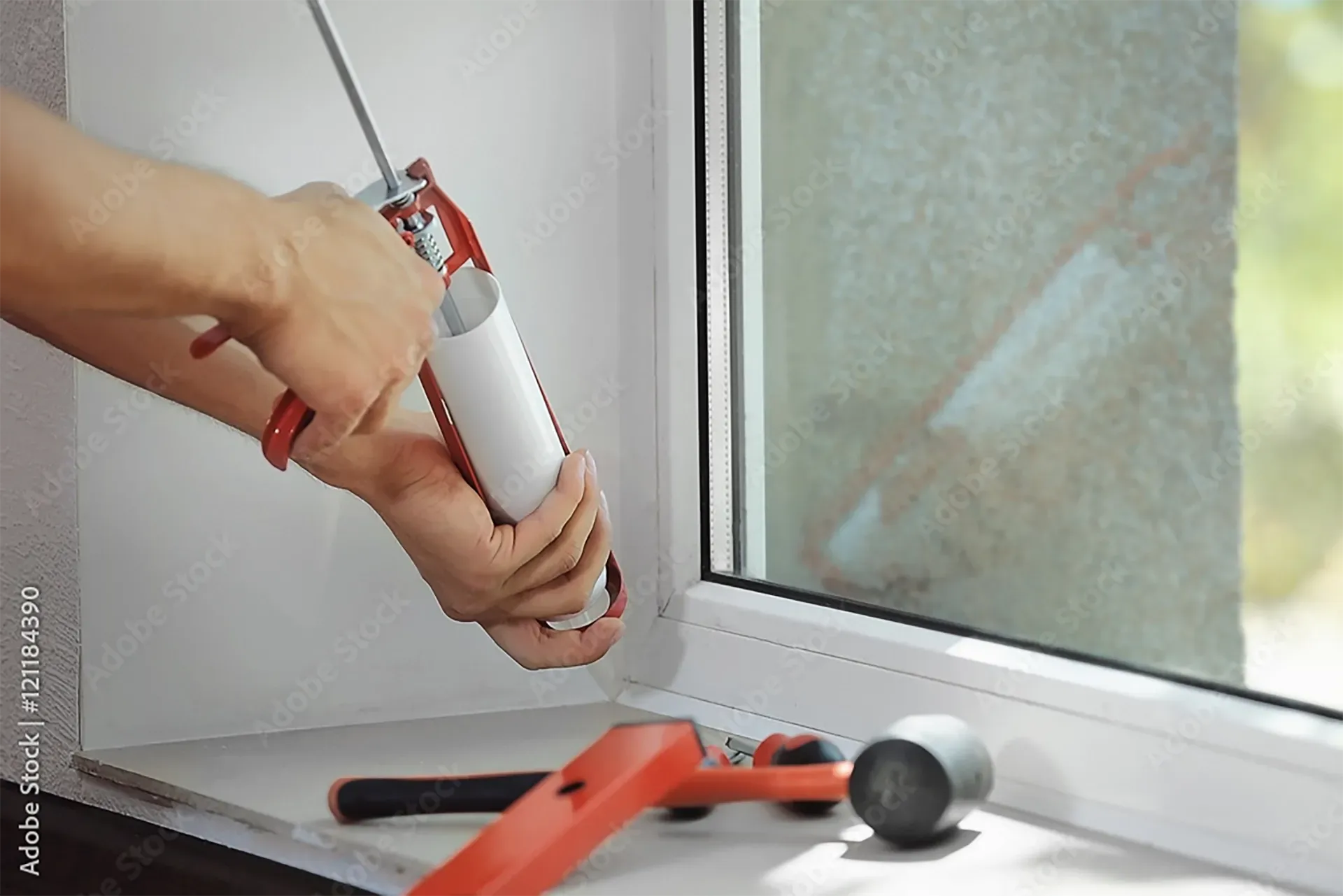 A person uses a red caulking gun to apply sealant along the edge of a white window frame, with tools resting nearby.