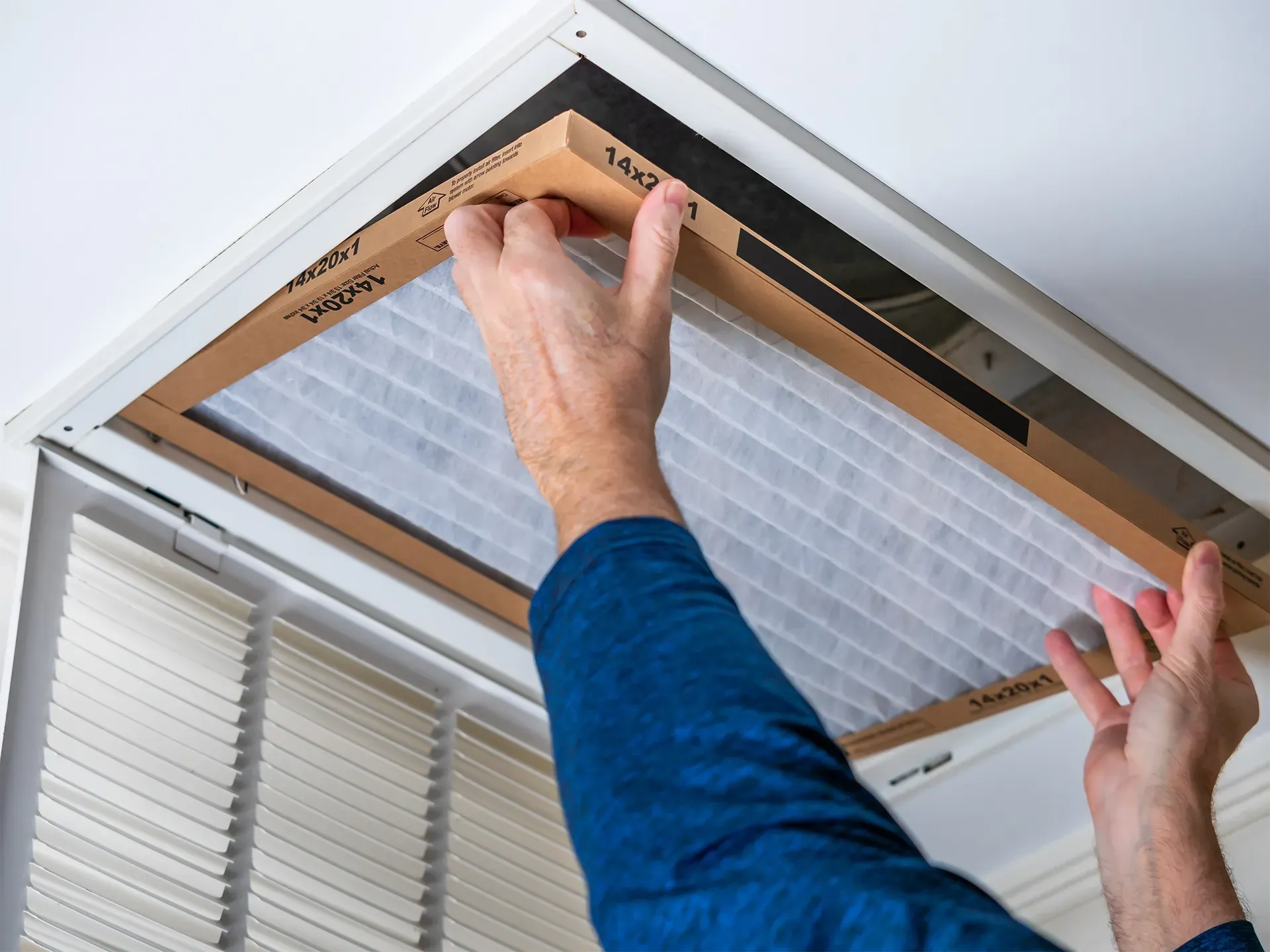 Hands placing a new rectangular air filter into a ceiling-mounted ventilation grate.