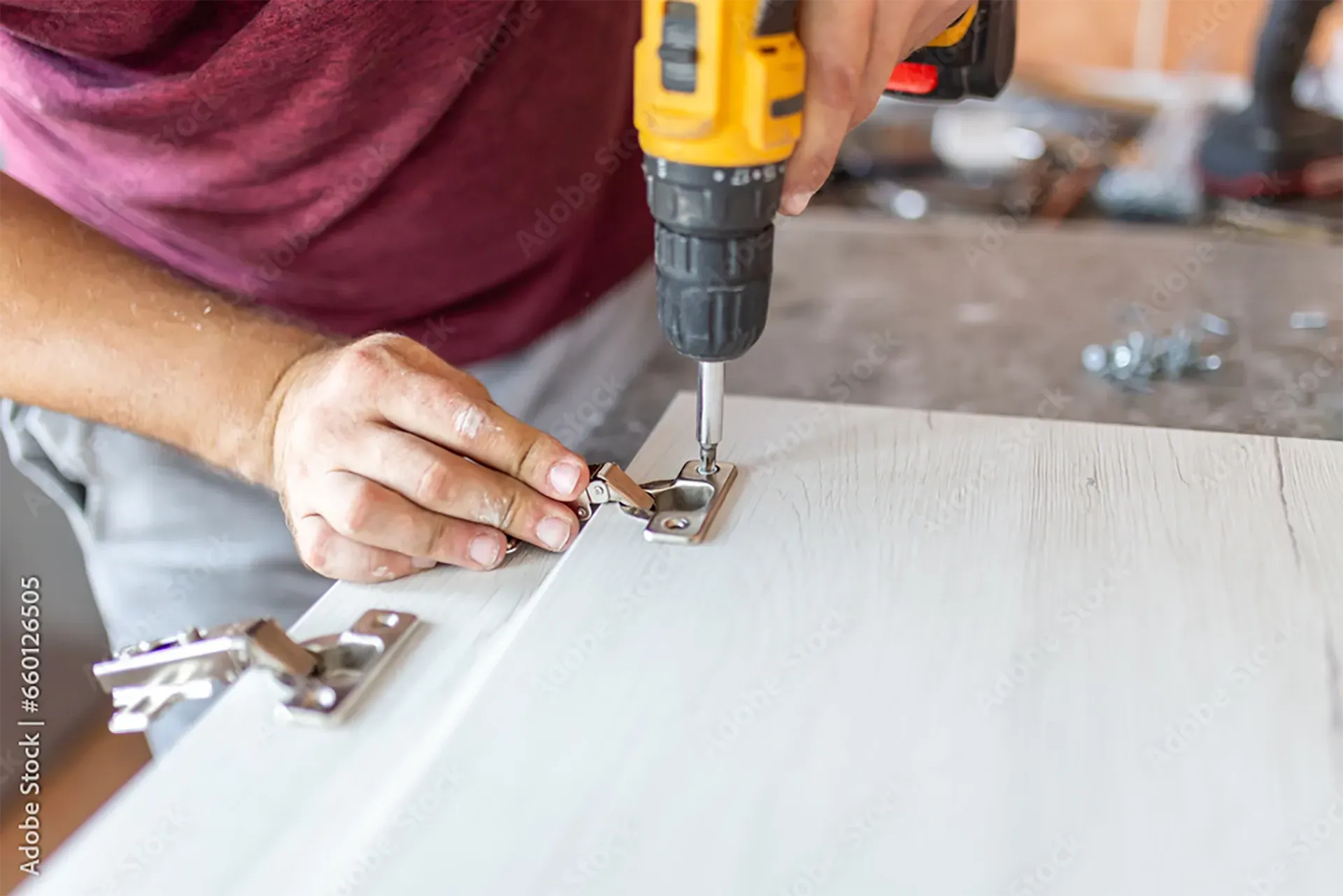 A person uses a yellow electric drill to attach a metal hinge to a light-colored wooden cabinet door.