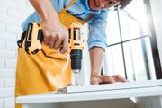 A person in a yellow apron uses a cordless power drill to install a metal hinge on a white wooden panel.