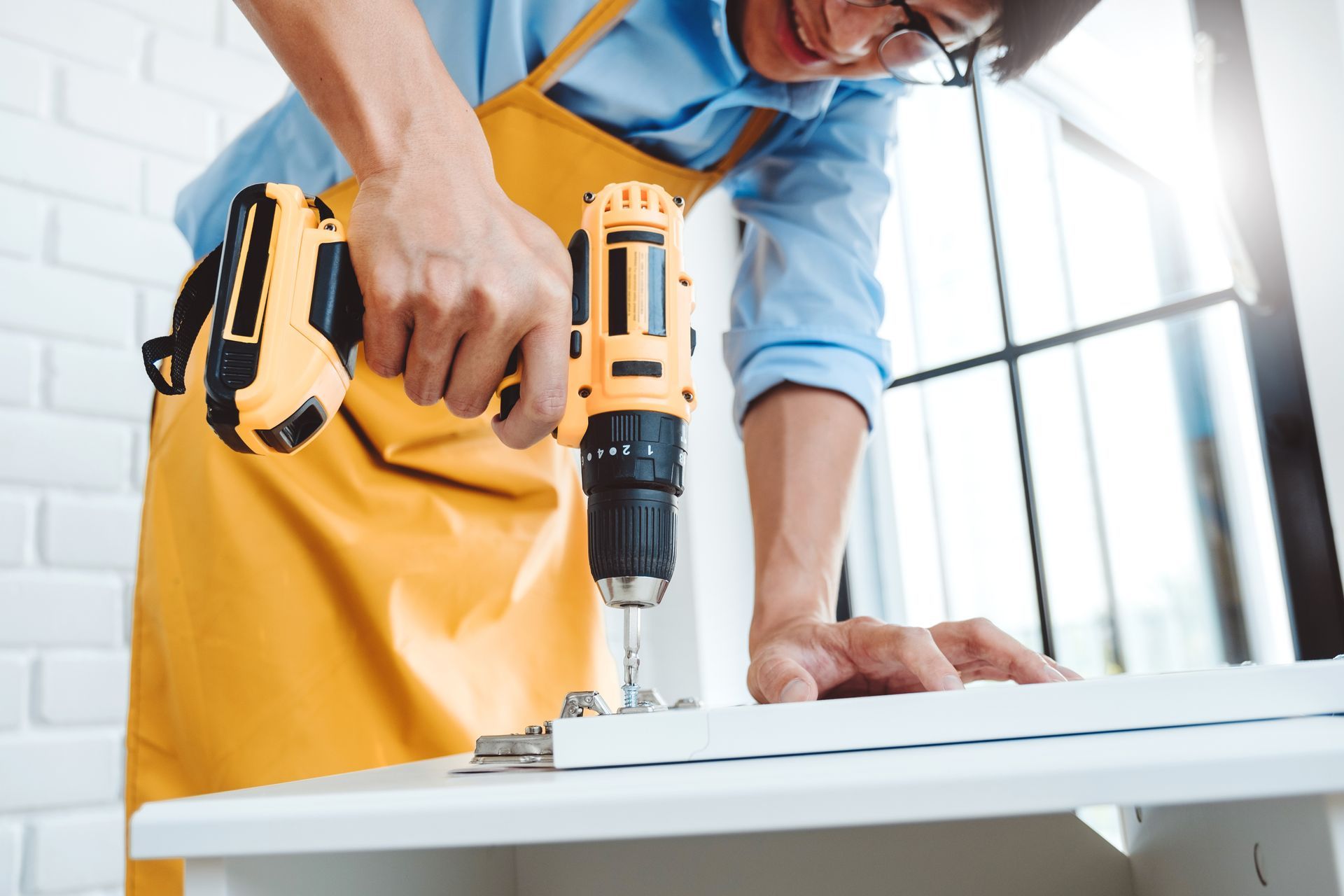 A person in a yellow apron uses a cordless power drill to install a metal hinge on a white wooden panel.