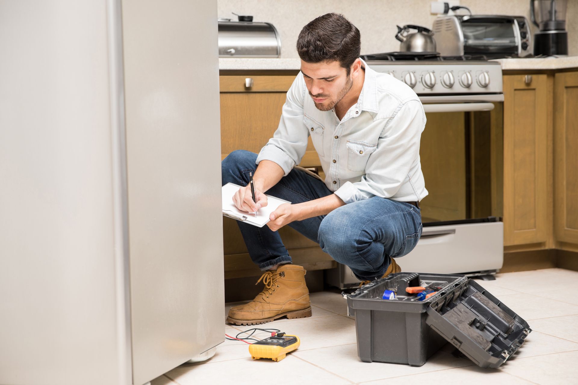 A technician kneels in a kitchen, writing on a notepad while inspecting a stainless steel refrigerator.