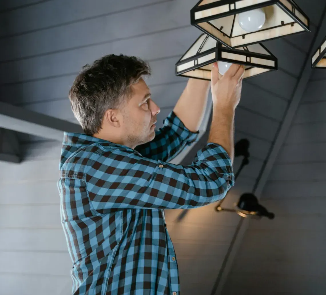 A person in a blue and black plaid shirt stands on a ladder, reaching up to change a lightbulb in a rustic pendant lamp.