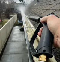 A person uses a pressure washer to clean a dirty rain gutter along the edge of a shingled roof.