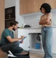 A technician kneels to inspect plumbing under a kitchen sink while a person stands nearby, looking on.