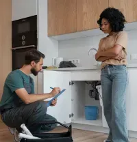A technician kneels to inspect plumbing under a kitchen sink while a person stands nearby, looking on.