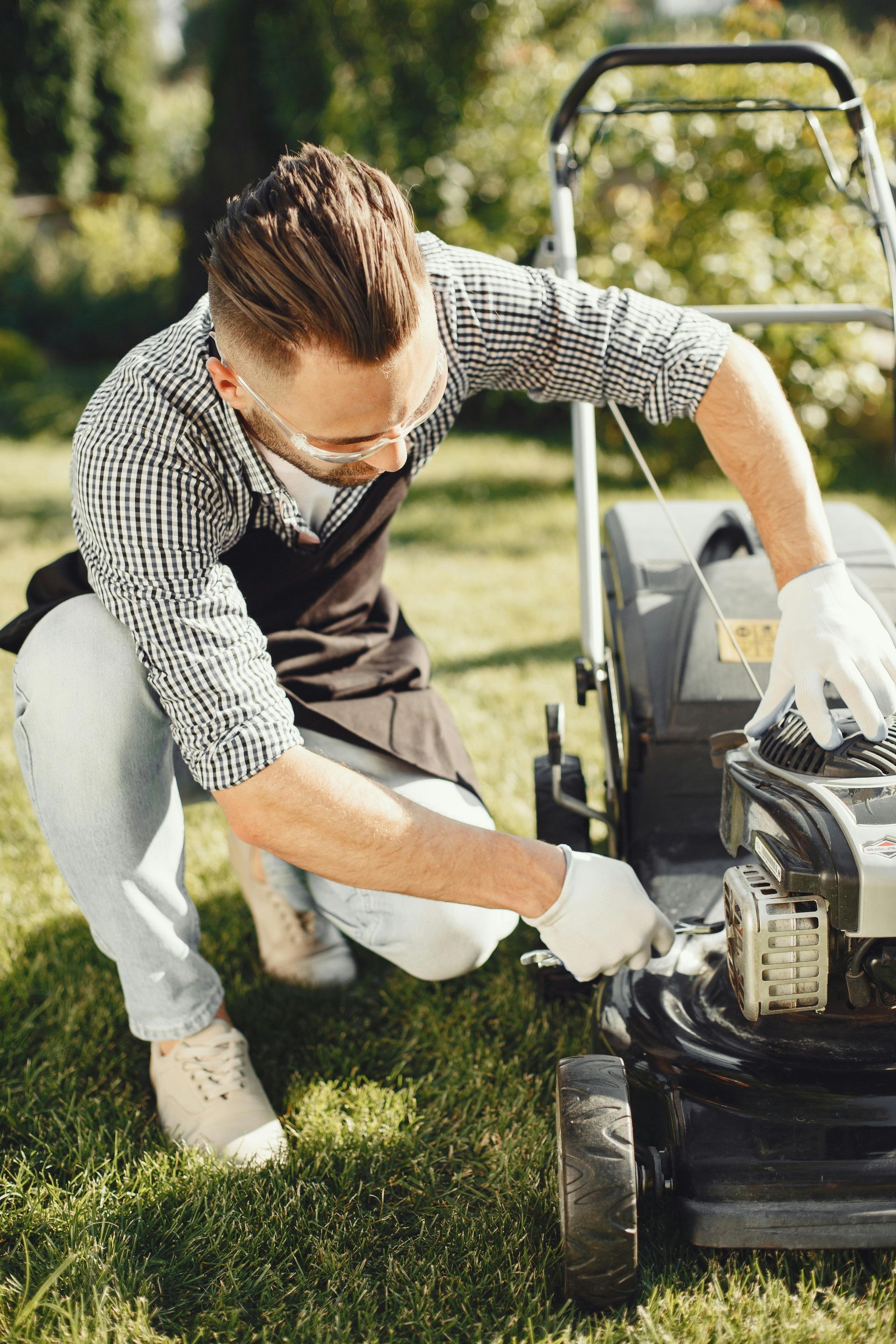 A person in an apron and gloves kneels on a lawn, repairing a black lawnmower outdoors on a sunny day.