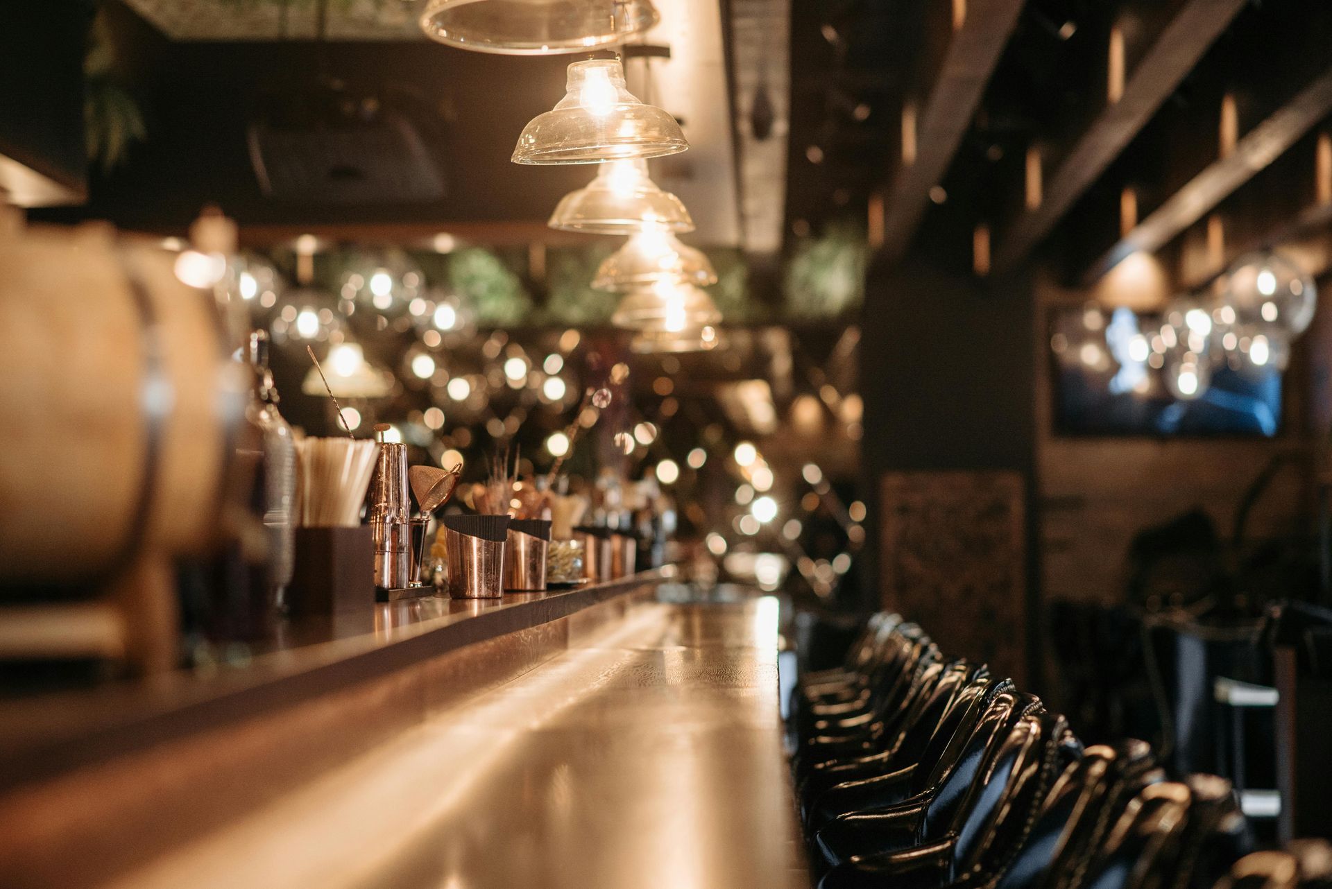An empty bar counter with warm, glowing pendant lights, wood surfaces, and a row of dark stools in a dim, upscale setting.