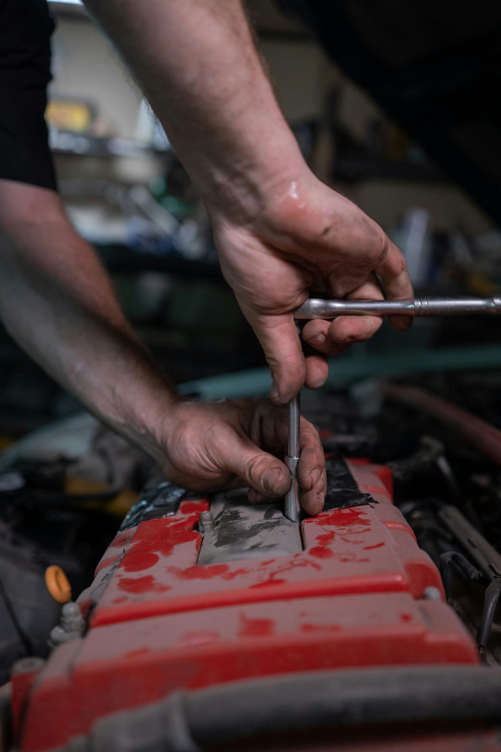 A mechanic uses a metal wrench and socket to work on a red car engine in a garage.