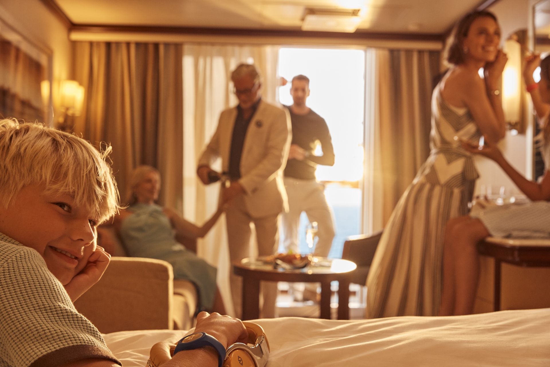 A family enjoys time together in their cruise stateroom at sunset, with warm light filling the room and an ocean view visible through the balcony doors.