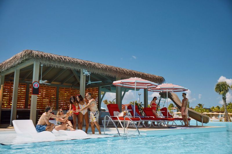 Poolside scene with people, bar, lounge chairs, umbrellas, and a slide under a clear blue sky.