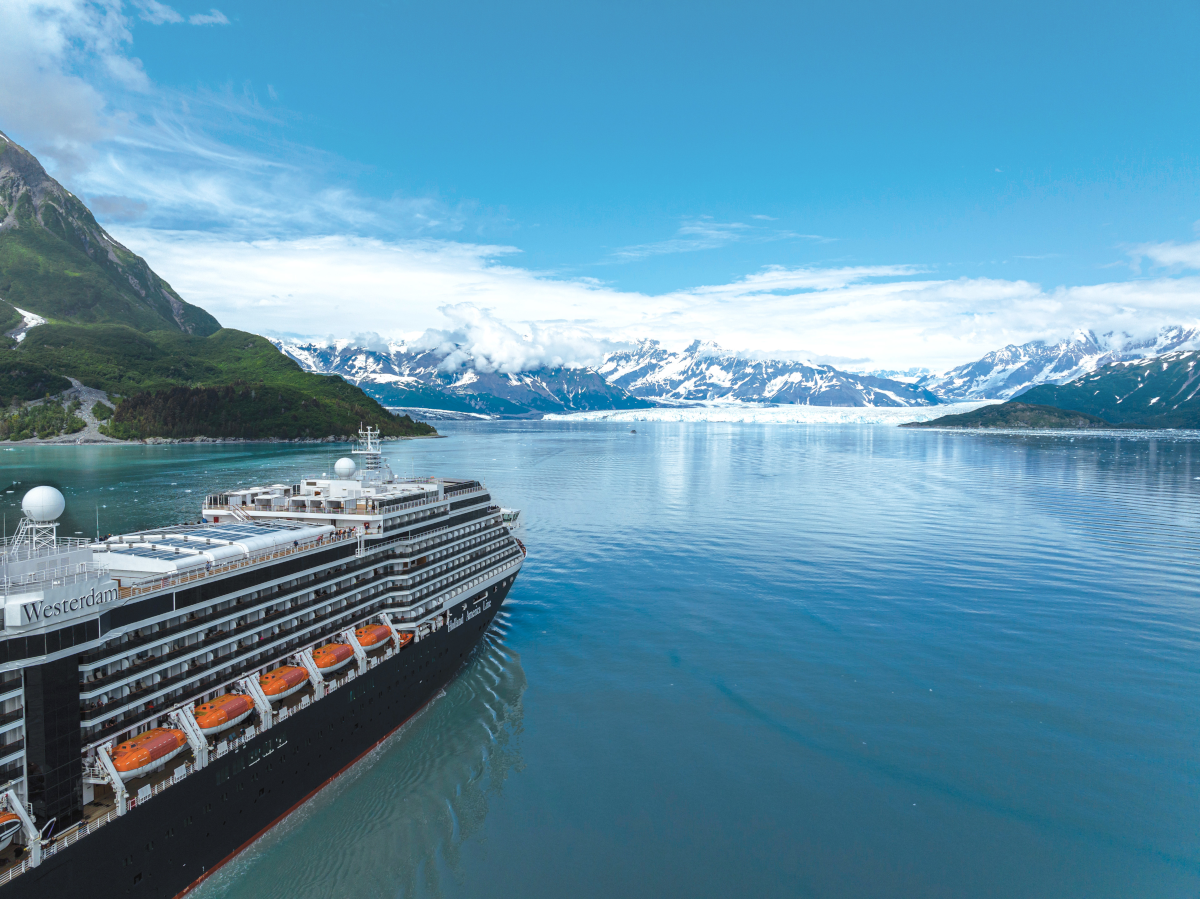 Cruise ship in a calm, blue bay with snow-capped mountains and green hills under a partly cloudy sky.
