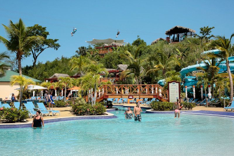 Swimming pool area with people, a bridge, slides, and buildings surrounded by palm trees under a blue sky.