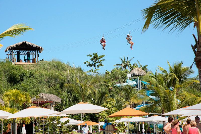 Two people zip-lining over a tropical resort with umbrellas, water slides, and a gazebo.