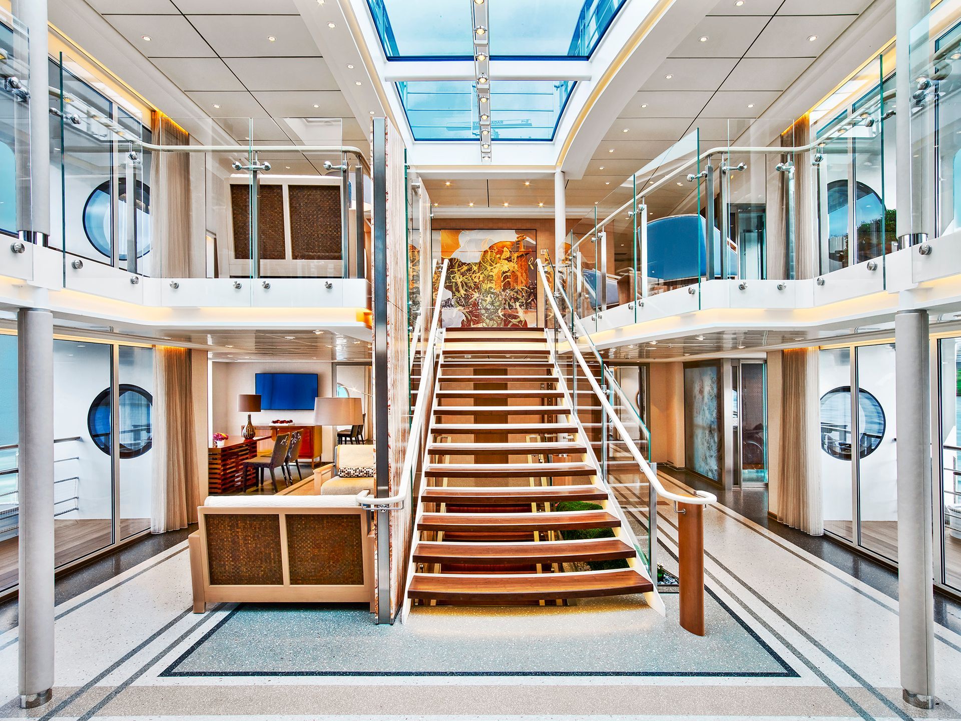 Interior view of a multi-story river cruise ship. Open staircase, glass railings, porthole windows, and a glass ceiling.