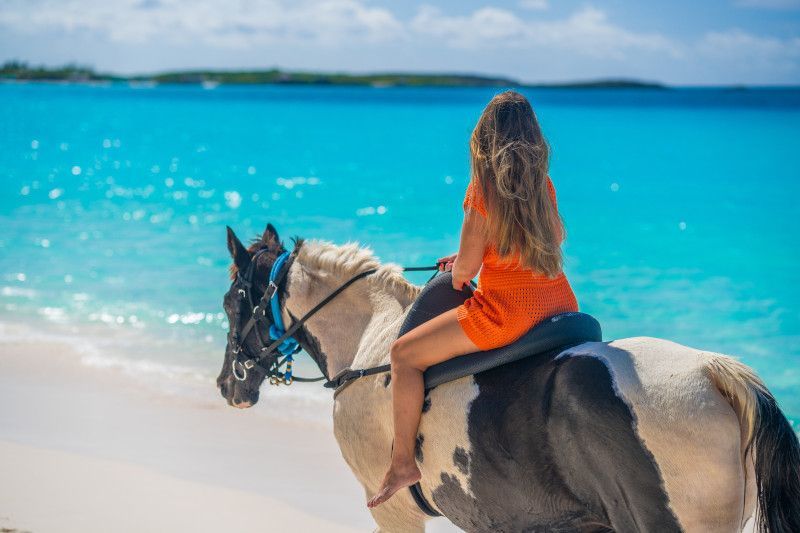 Woman riding a painted horse on a beach, turquoise water and blue sky in the background.