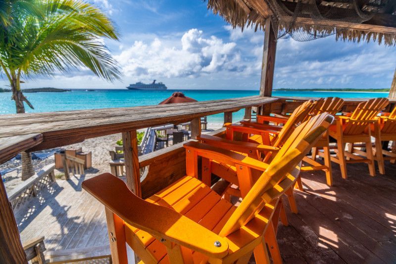Beach bar with orange chairs, thatched roof, and ocean view with a cruise ship in the distance.