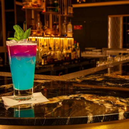 Cocktail on a marble bar with a dark background. The drink is blue, purple, and layered with mint garnish.