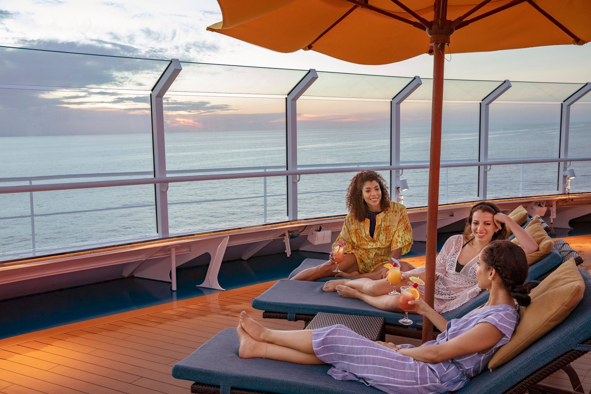 Three women relaxing on deck chairs under an umbrella, ocean view, enjoying drinks.