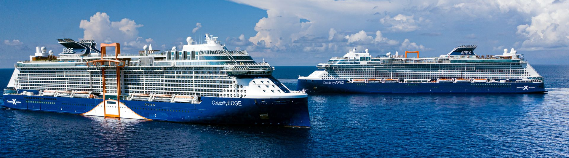 Two blue cruise ships, sisters Edge and Apex, in the ocean under a cloudy sky.
