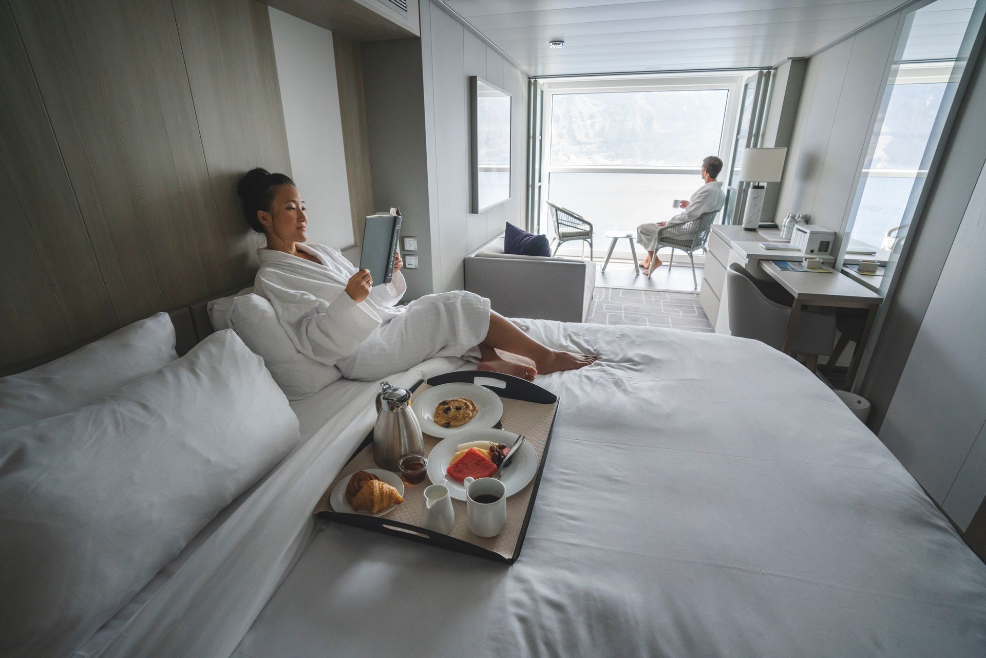 Woman in robe reads in bed, breakfast tray present. Man on balcony. Modern, bright cabin.