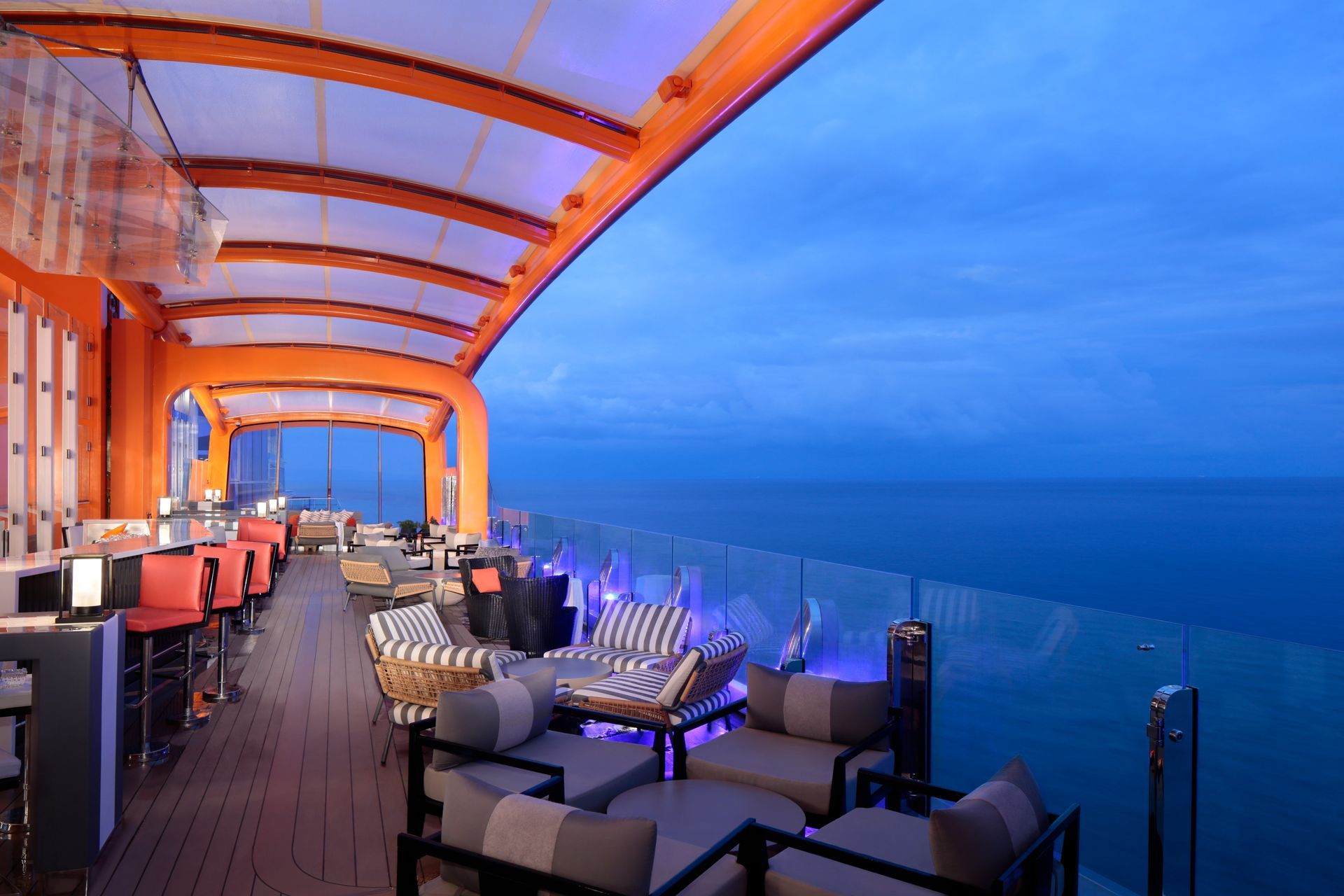 A rooftop bar at dusk overlooking the ocean. Orange accents, seating, and a clear blue sky.