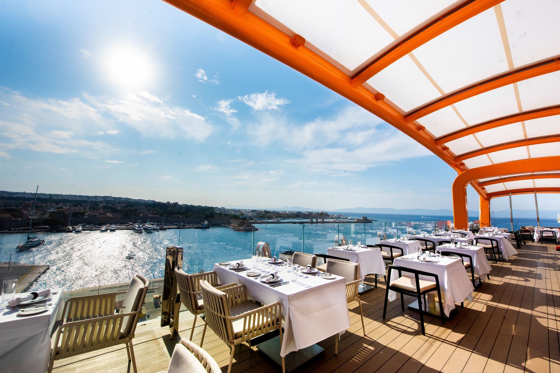 Rooftop restaurant with tables set for dining, overlooking a blue sea and coastline under a bright sky.