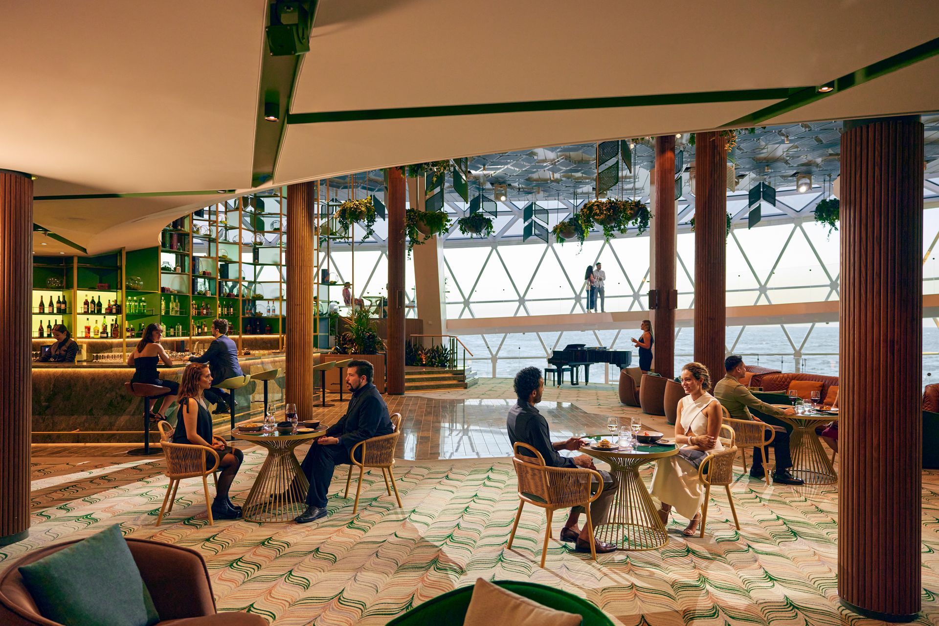 Interior of a modern restaurant with tables, people, and a bar overlooking the ocean. Green and neutral tones are dominant.