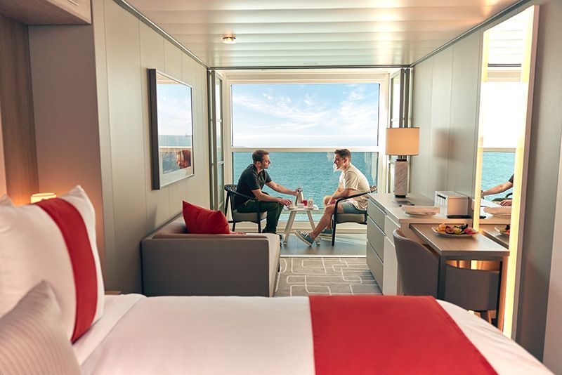 Cruise ship stateroom with two people on balcony, ocean view. Interior has bed, couch, and desk.
