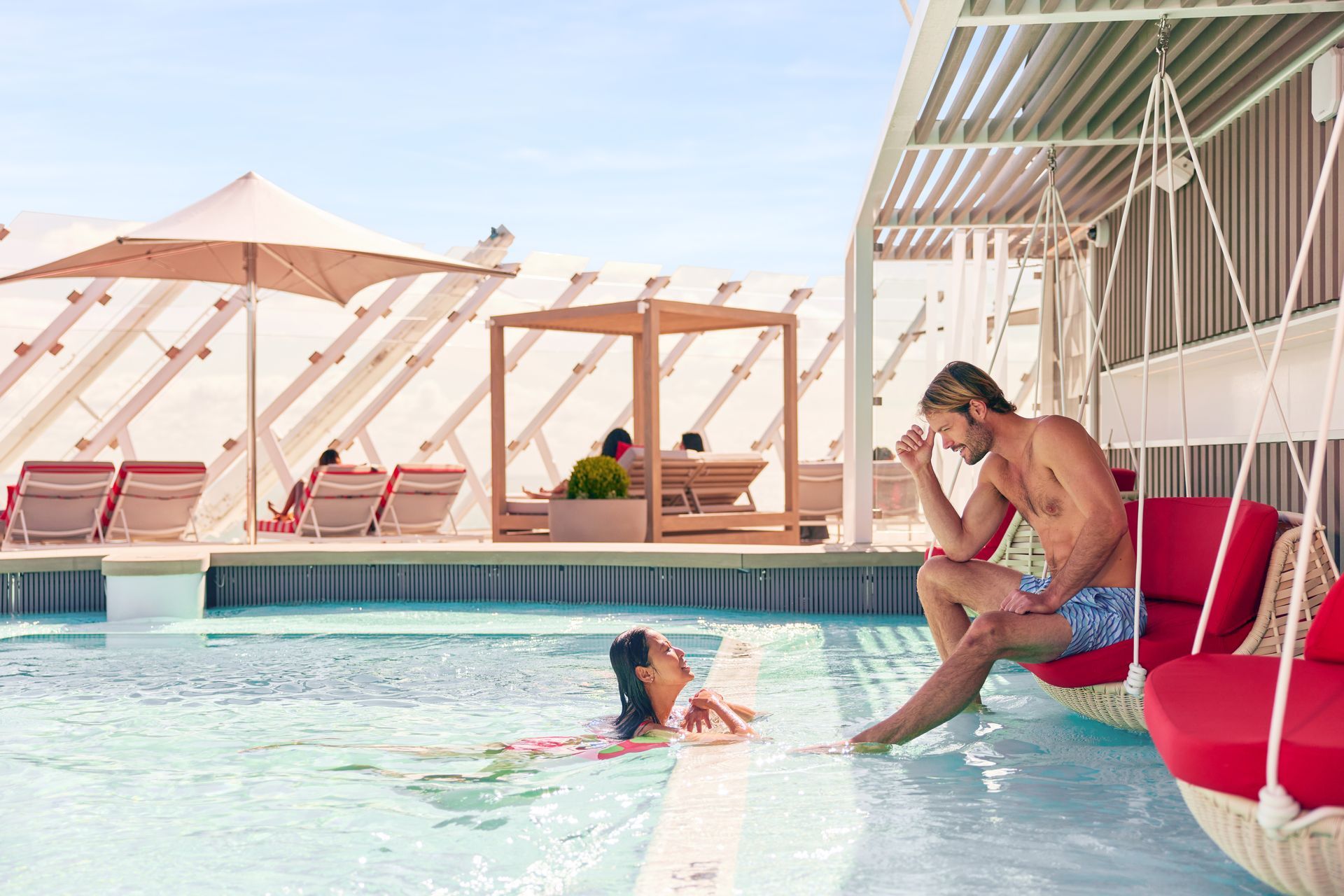Man sits by pool, woman swims. Red cushions, white structure, sunny outdoor setting.
