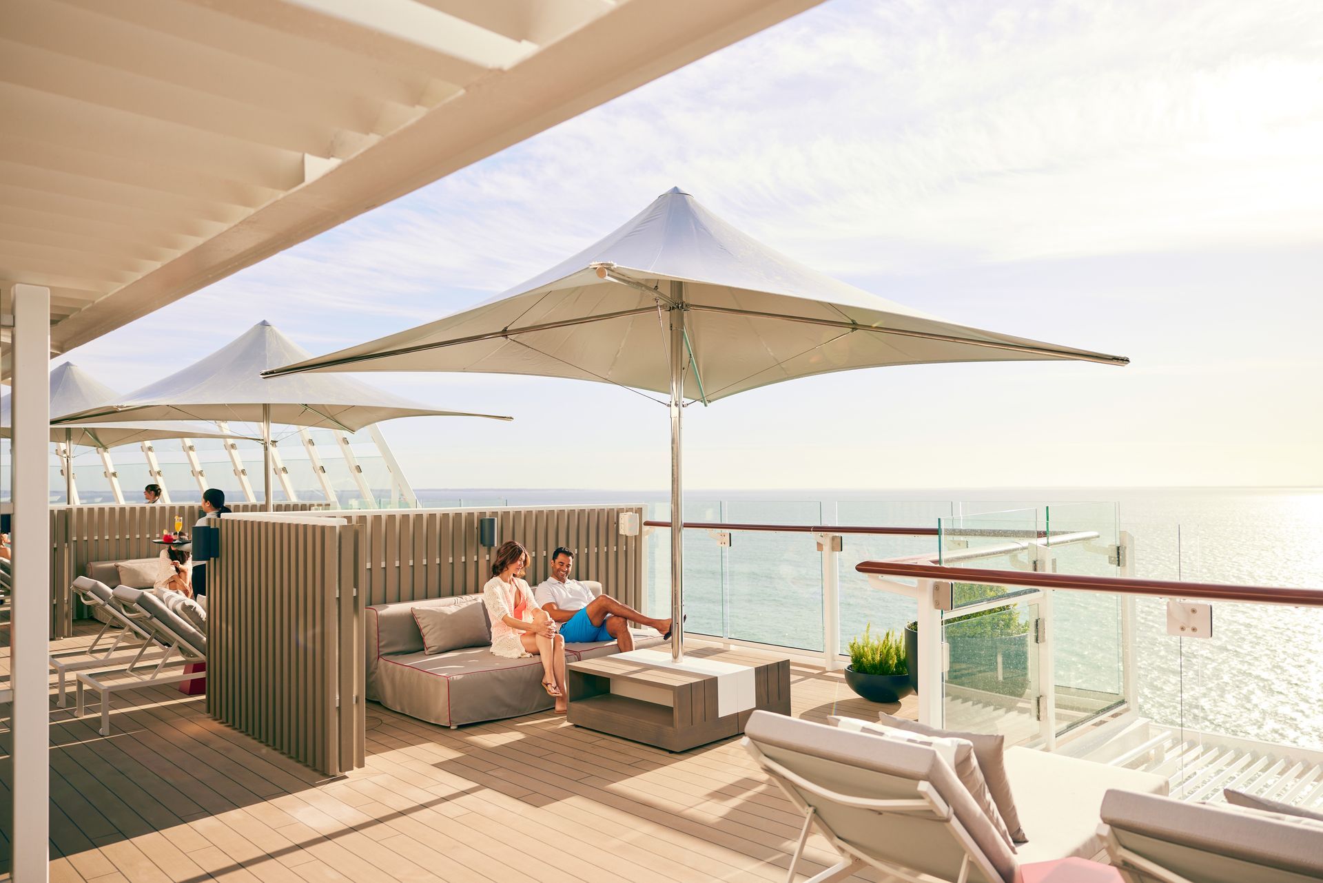 Couple relaxing on outdoor deck, beneath a large umbrella, overlooking the ocean.