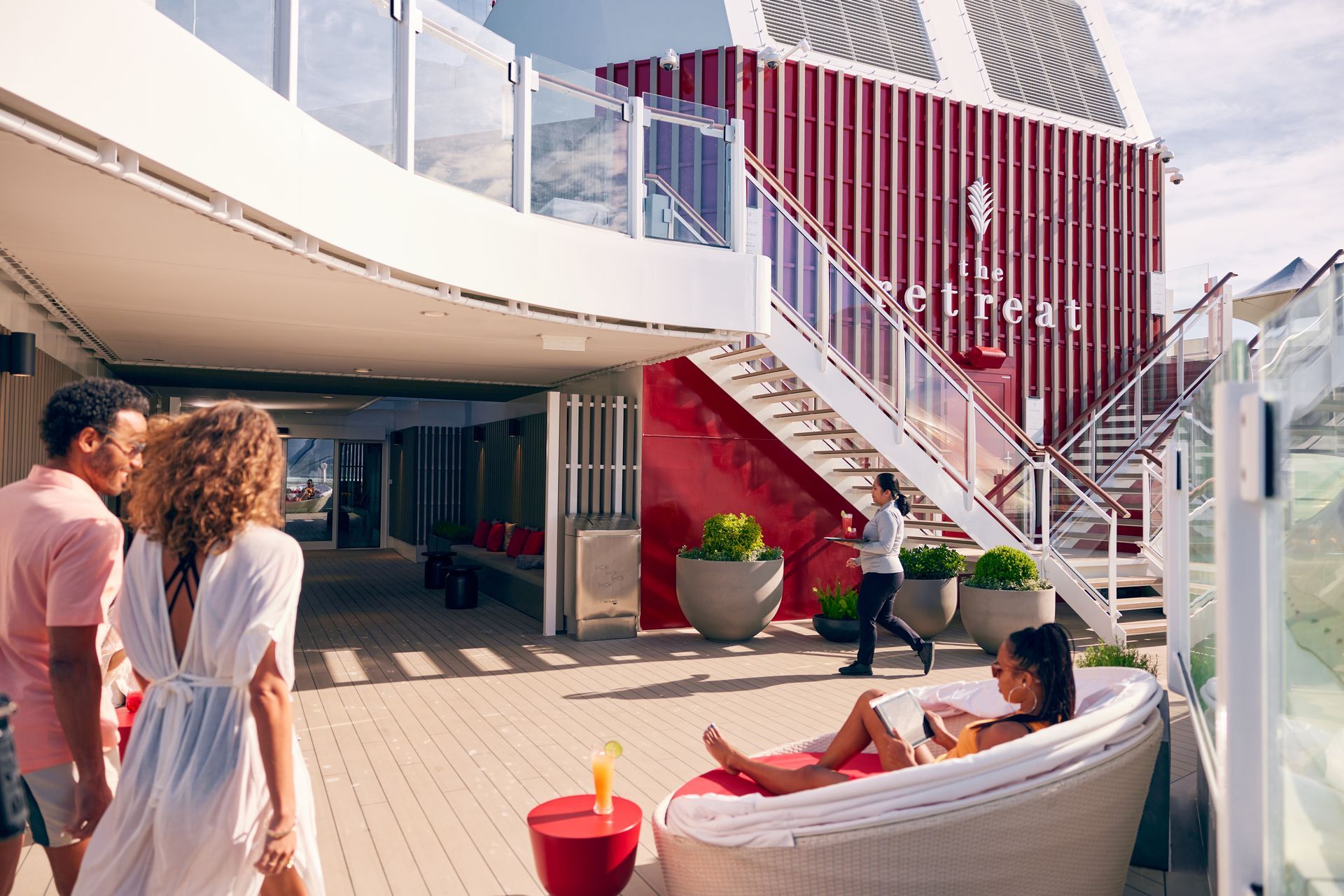 People on a cruise ship deck. One person reclines on a white circular lounge chair, others walk, red accents.