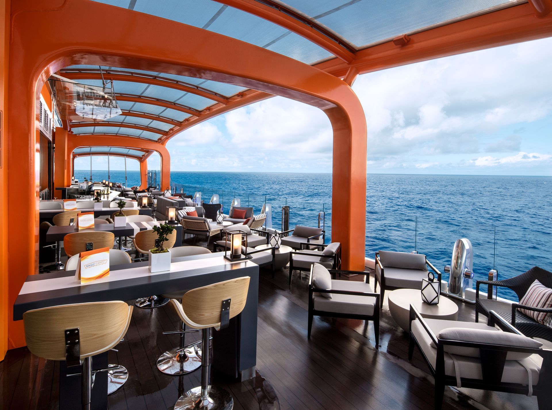 Outdoor cruise ship lounge overlooking the ocean, with orange frame, tables, chairs, and bar.