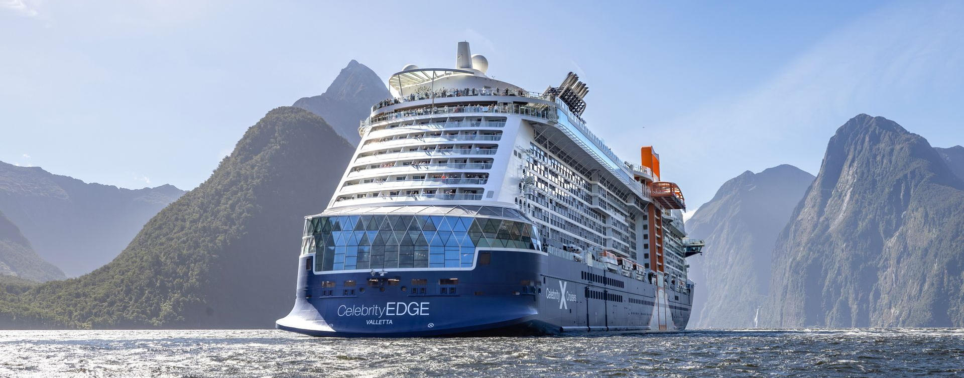 A large cruise ship sailing in front of large, forested mountains in a sunny setting.