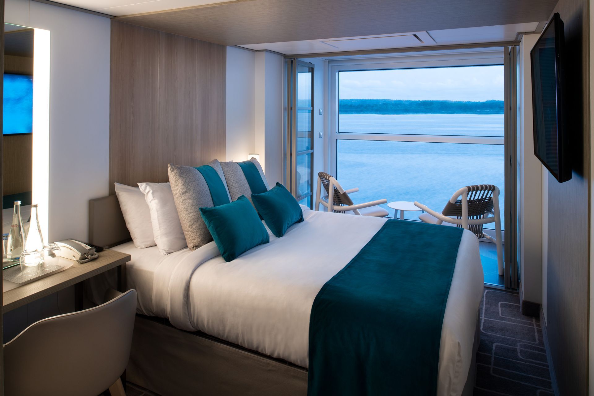 Bedroom on a cruise ship with a bed, small balcony with ocean view, and a TV on the wall.