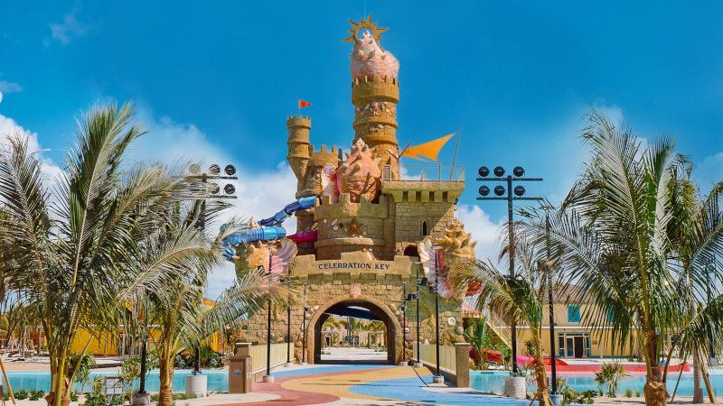  A massive, elaborate sandcastle-themed water park structure with slides and a central archway entrance stands under a blue sky at Celebration Key.