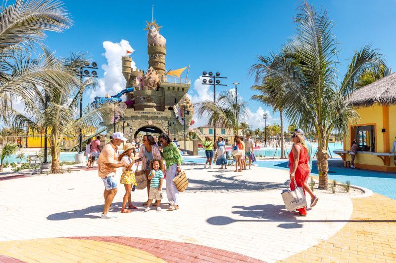 People at a sunny resort near a castle-like structure, palm trees, and pools.